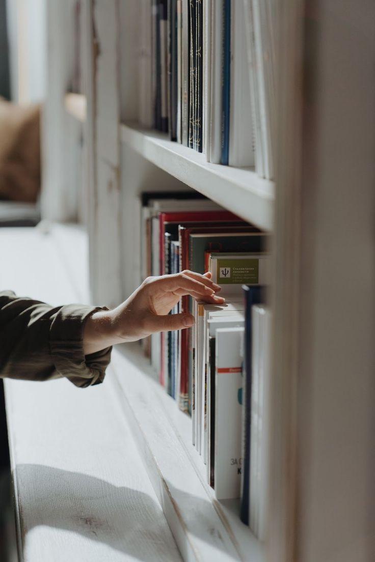 A hand reaching for a book on a white library shelf, lit by soft window light